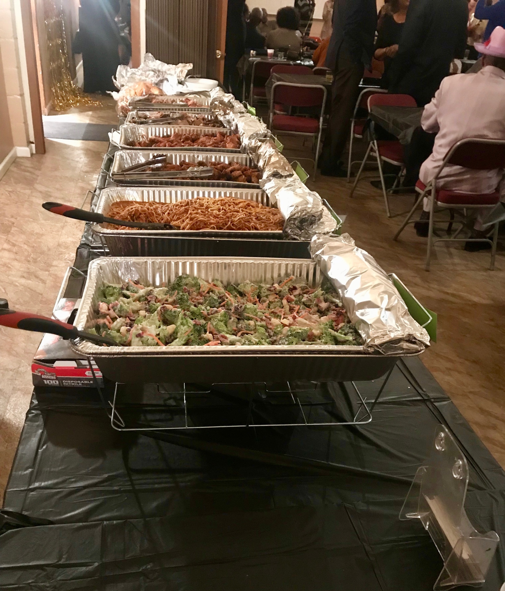 Long indoor buffet line with chafing pans of spaghetti, wings, and broccoli salad